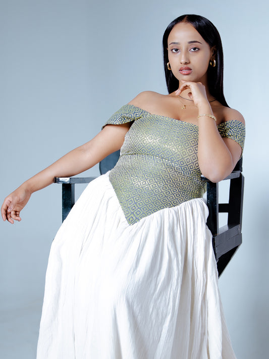 A woman posing in a white maxi Habesha Dress with a textured metallic green off-the-shoulder bodice, seated on a black chair against a light gray background.