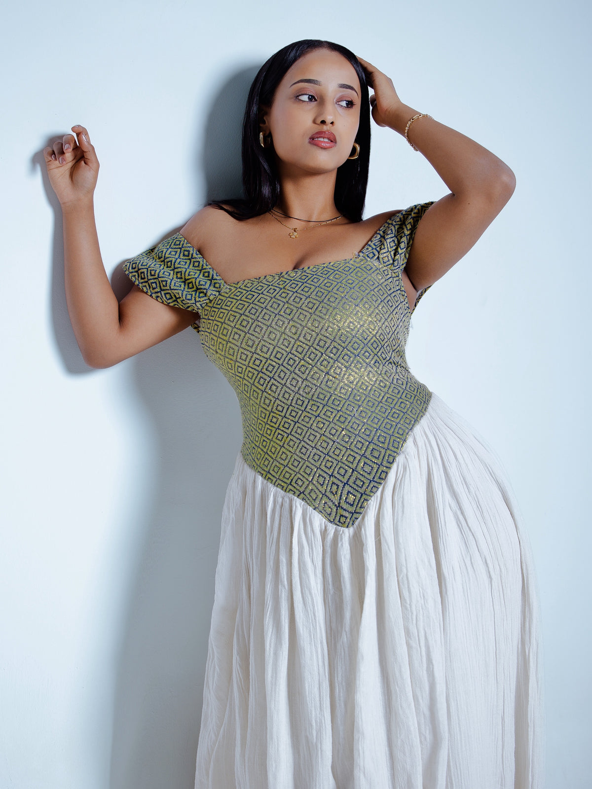 A woman posing in a green and gold patterned off-the-shoulder top with a long white pleated Habesha Dress, standing against a light blue wall.
