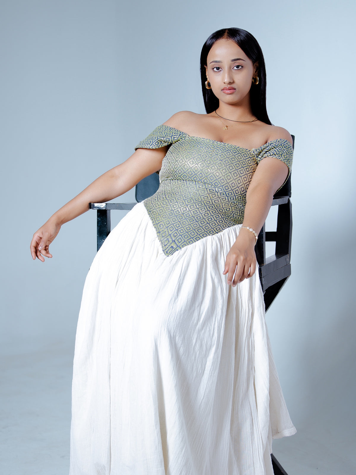 A woman wearing a shimmering off-the-shoulder Habesha Dress with a textured metallic finish and a long flowing white skirt, seated on a black chair against a light gray studio background.