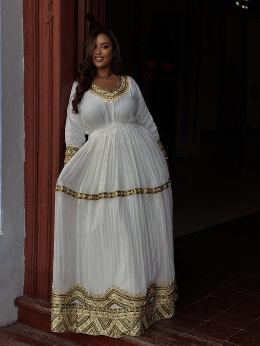 A woman modeling a white long-sleeved maxi Habesha Kemis with ornate gold embroidery, standing in a dimly lit hall with chandeliers in the background.