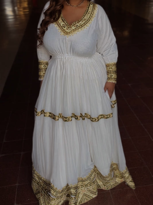 A woman modeling a white long-sleeved maxi Habesha Kemis with ornate gold embroidery, standing in a dimly lit hall with chandeliers in the background.