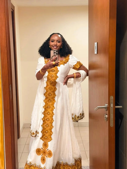 A woman takes a mirror selfie wearing a white traditional Ethiopian Dress with prominent gold embroidery and patterns. The photo is taken in an indoor setting with wooden doors on both sides.