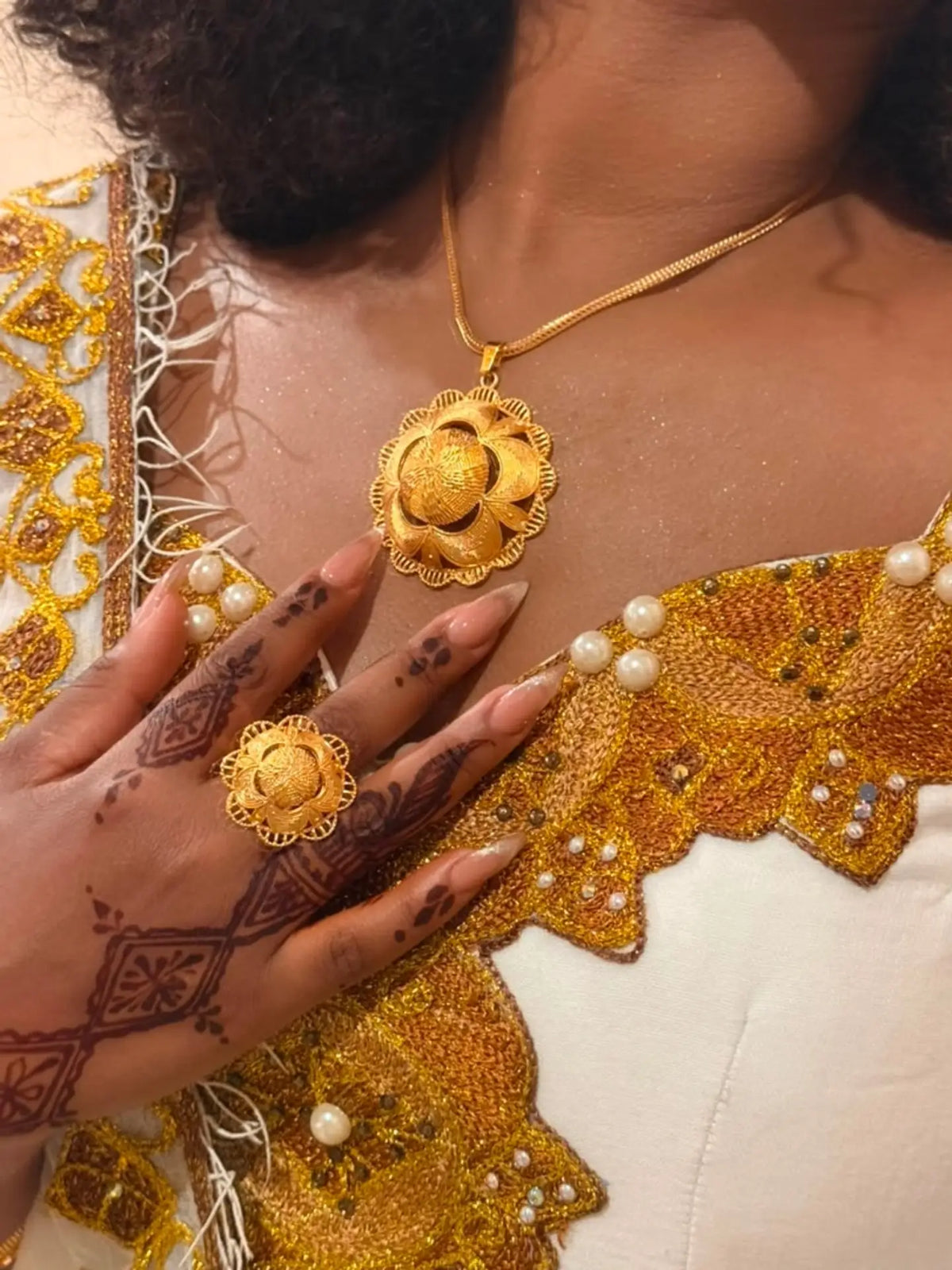 A close-up of a person wearing a gold necklace with a flower-shaped pendant and a matching gold ring, styled with a white and gold embroidered traditional Ethiopian Dress.