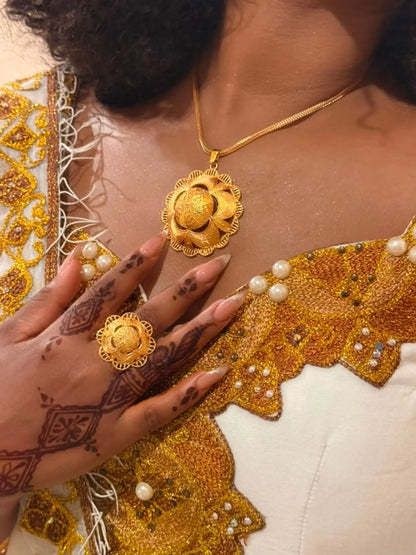 A close-up of a person wearing a gold necklace with a flower-shaped pendant and a matching gold ring, styled with a white and gold embroidered traditional Ethiopian Dress.