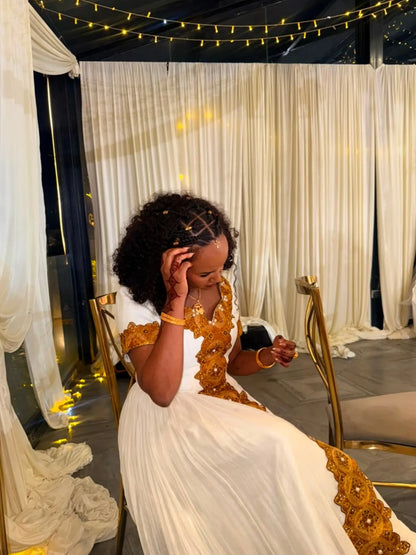 A woman seated indoors wearing a white Ethiopian Dress with ornate gold embroidery and gold jewelry, surrounded by draped white fabric, string lights, and gold chairs.