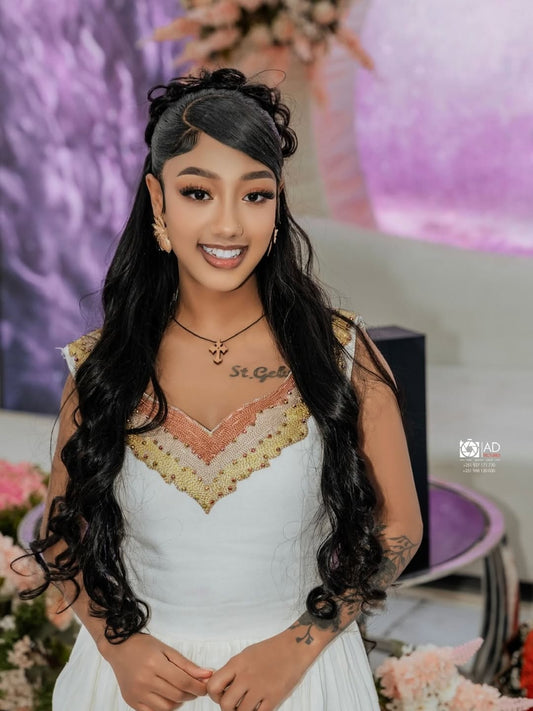 Woman in a white Gold Eritrean Dress with floral details, standing in front of a blurred decorative background.