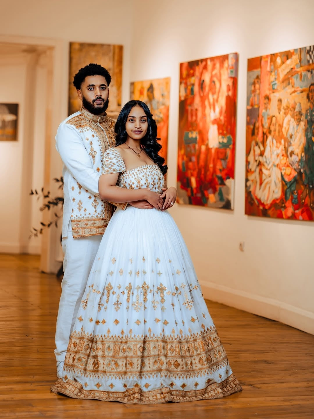 Couple in traditional Gold Unique Ethiopian Wedding Matching Couple Set for Her and Him –  standing in an art gallery with colorful paintings on the wall.
