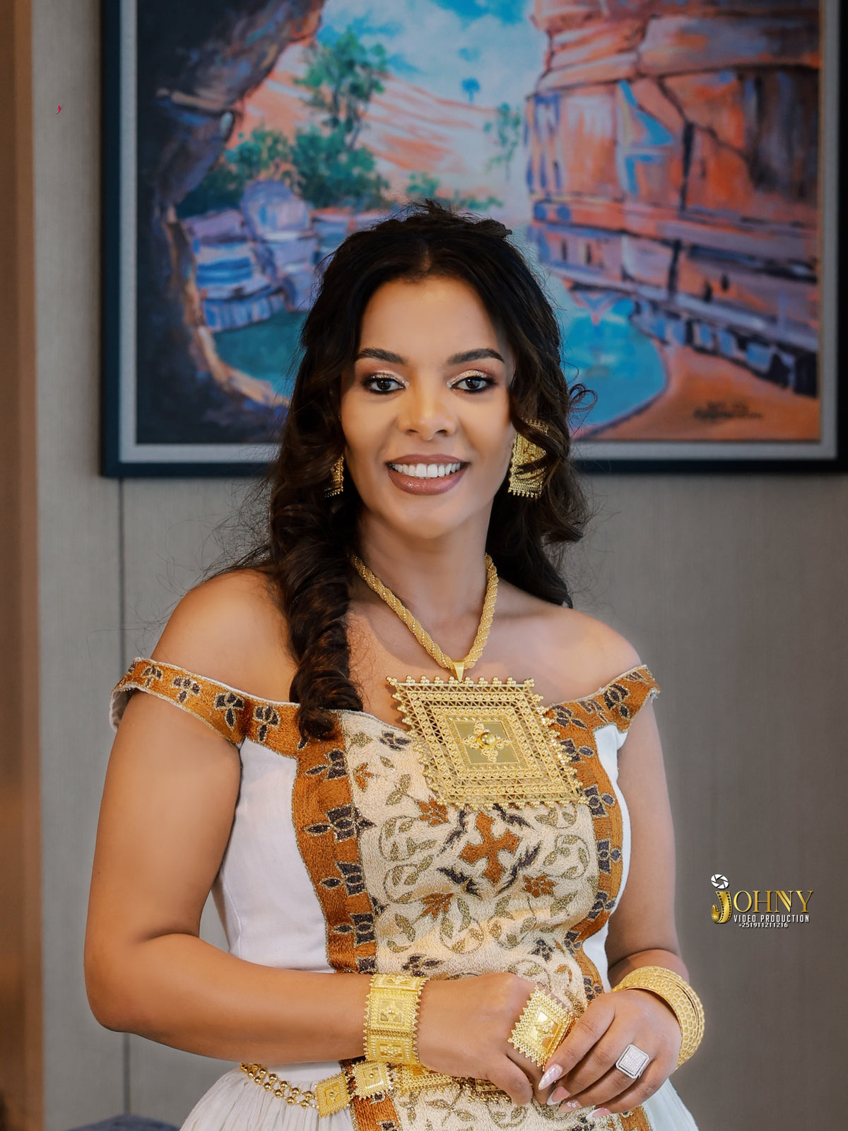 A woman models a traditional Ethiopian white Habesha Kemis with gold embroidery and a decorative gold sash, accessorized with gold jewelry in an indoor setting.