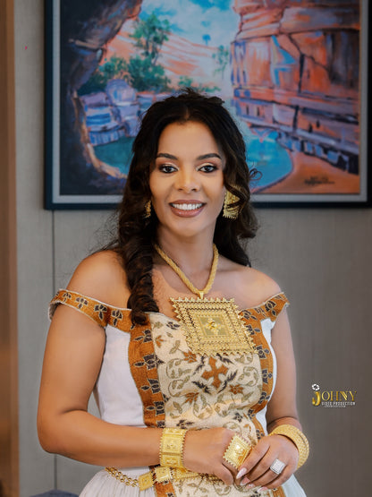 A woman models a traditional Ethiopian white Habesha Kemis with gold embroidery and a decorative gold sash, accessorized with gold jewelry in an indoor setting.