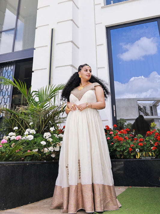 A woman wearing a cream and beige floor-length Habesha Kemis with gold embellishments, standing outdoors in front of a modern white building with large windows and flower beds.