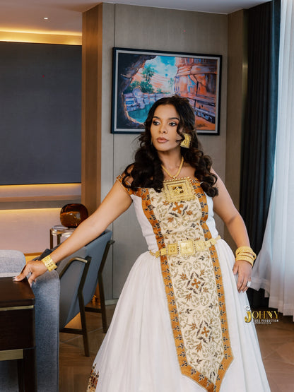 A woman models a traditional Ethiopian white Habesha Kemis with gold embroidery and a decorative gold sash, accessorized with gold jewelry in an indoor setting.