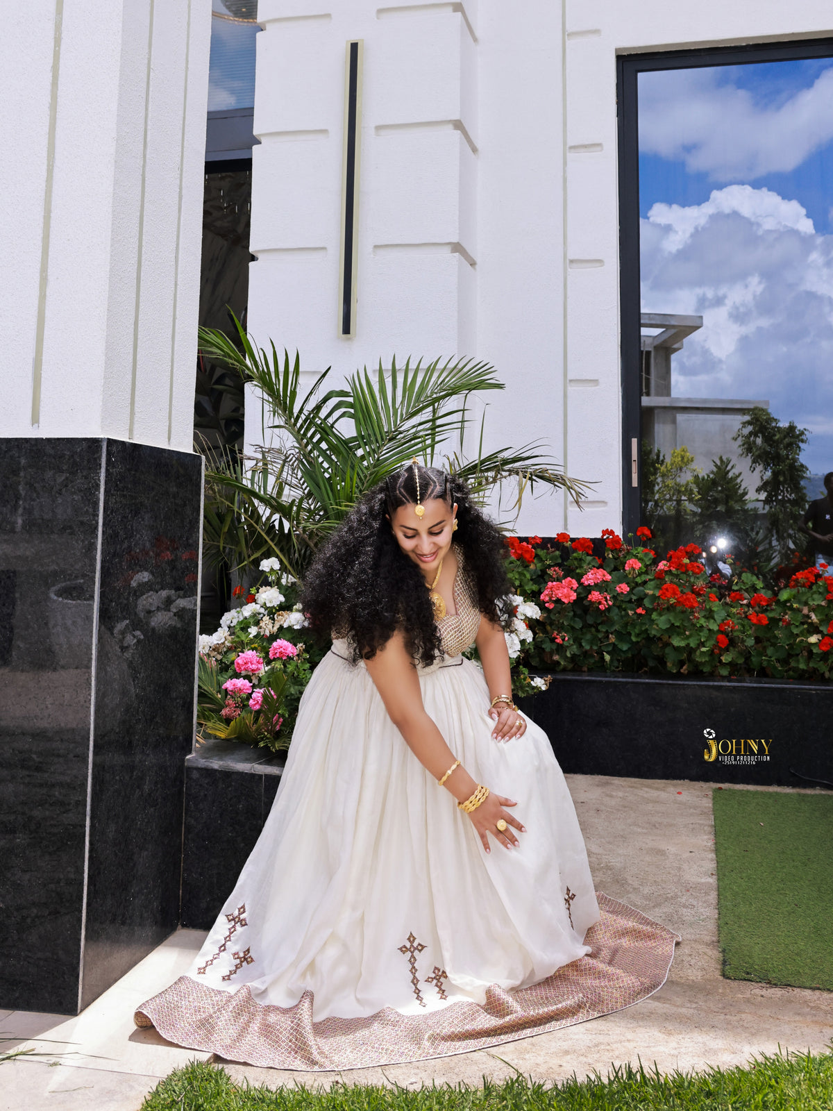 A woman wearing a white sleeveless Habesha Kemis with gold embroidery and a pinkish-beige hem, standing outdoors in front of a modern white building with flowers and greenery.