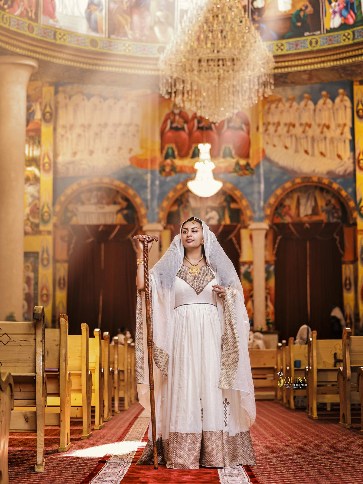 A woman stands in the center aisle of an ornate church interior, wearing a long white and gold Habesha Kemis with a sheer veil and holding a wooden staff. The background features painted religious murals, wooden pews, and a large hanging chandelier.
