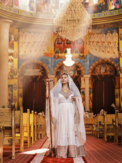 A woman stands in the center aisle of an ornate church interior, wearing a long white and gold Habesha Kemis with a sheer veil and holding a wooden staff. The background features painted religious murals, wooden pews, and a large hanging chandelier.