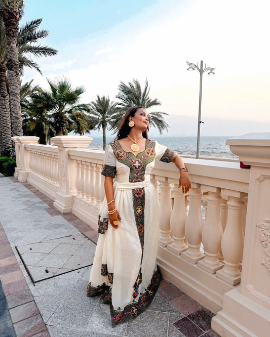 A woman wearing a traditional embroidered Ethiopian dress stands on a seaside terrace with palm trees and a balustrade railing behind her.