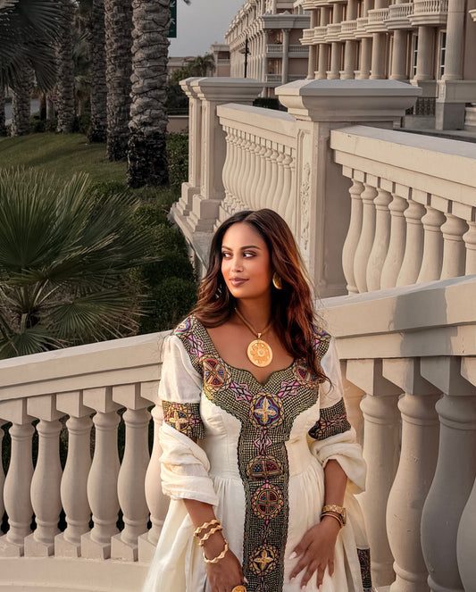 A woman wearing a white traditional Ethiopian dress with ornate multicolor embroidered details and gold jewelry, standing on a white stone terrace with palm trees and a grand building in the background.