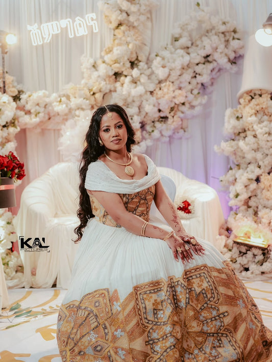Woman in a white and gold Gold Ethiopian Wedding Kemis sitting in a decorated room with flowers.