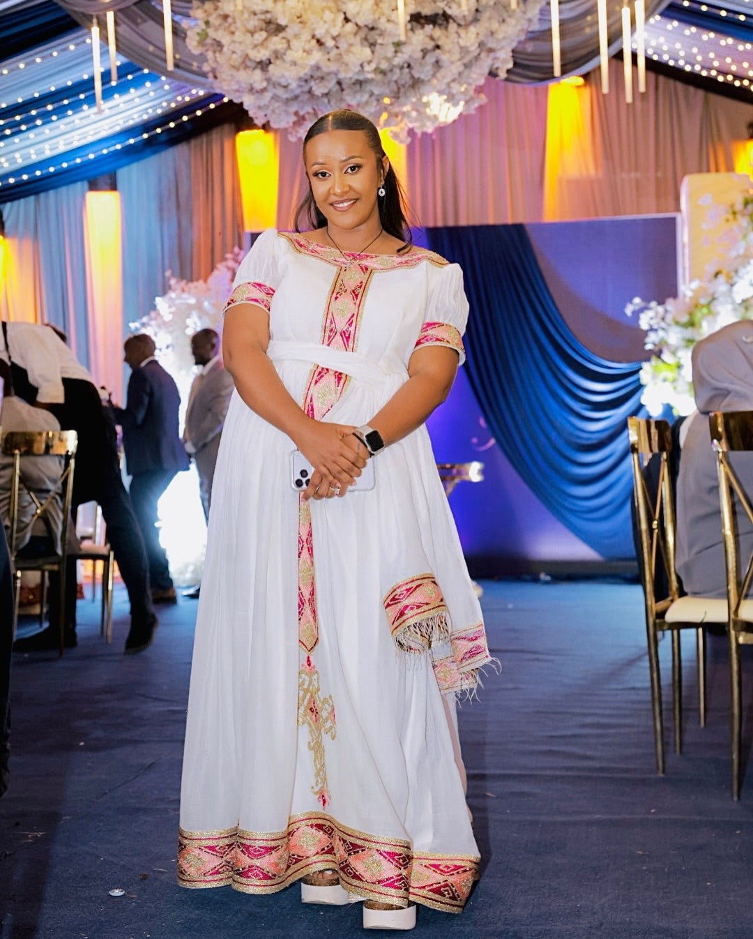 Woman in a white and pink traditional kemis standing in an event hall with decorations.