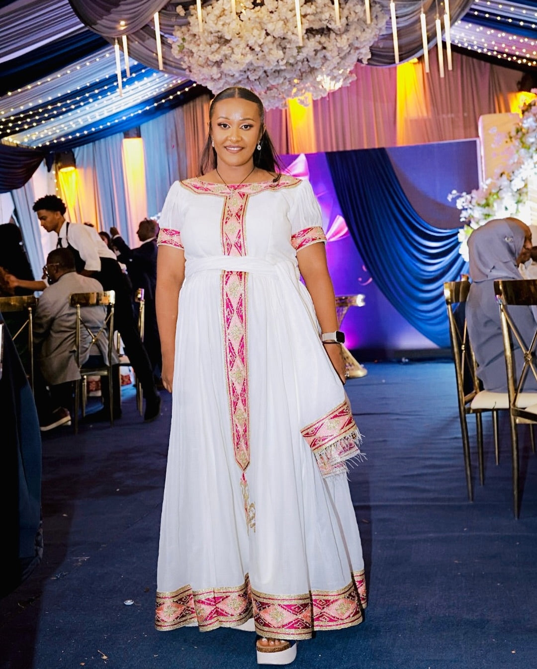 Woman in a white habesha kemis with pink and gold accents standing in an event hall.