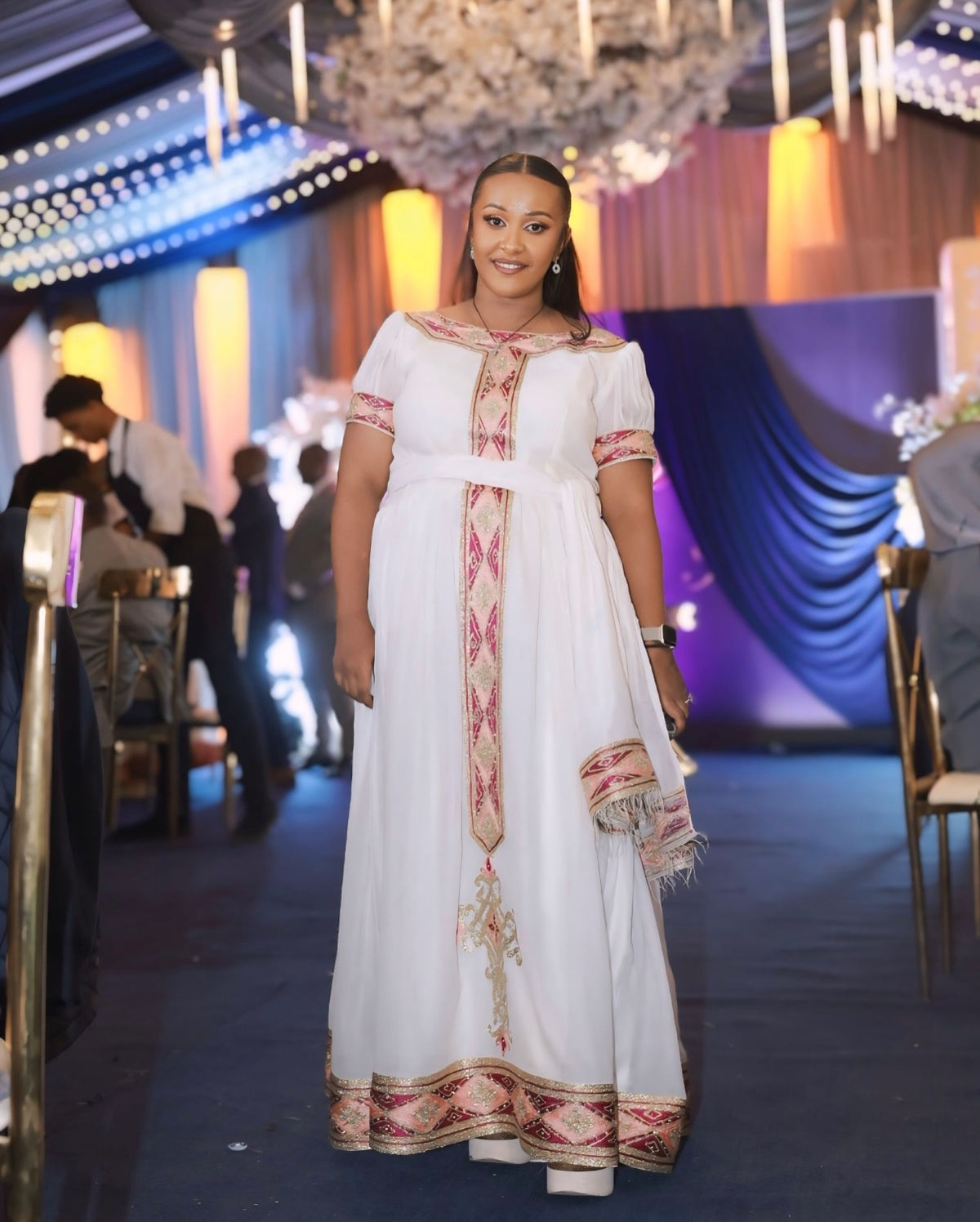 Woman in a white Habesha Kemis with red and gold patterns standing in an indoor setting with decorative lights.