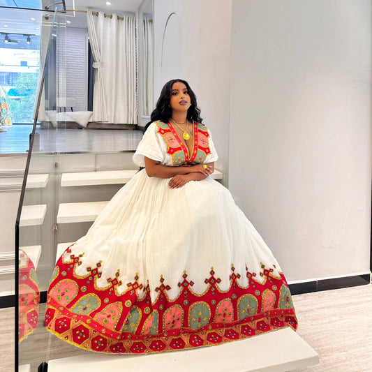 A woman wearing a traditional white habesha Wedding dress with vibrant red, gold, and green embroidery, posing on white stairs inside a modern building.