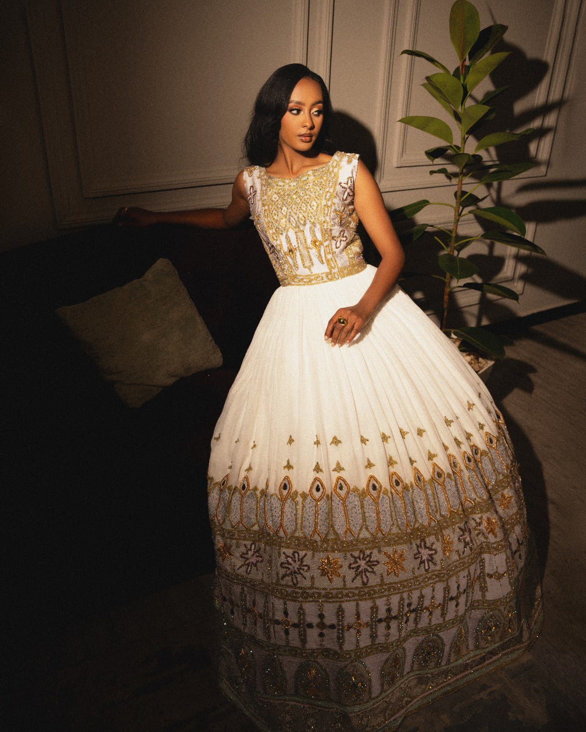 A woman models a voluminous white evening Habesha Wedding Dress  with intricate gold and silver embroidery, standing in a dimly lit interior next to a potted plant.