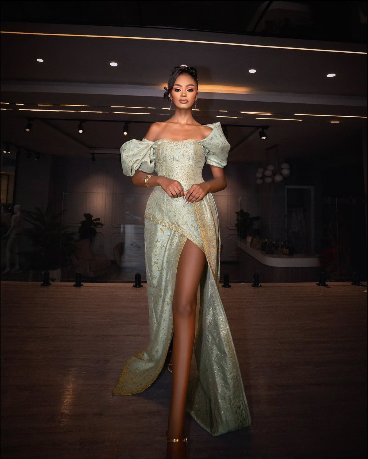 A woman models a floor-length, off-the-shoulder Habesha Wedding Dress with a high thigh slit in a dimly lit indoor venue. The dress has a shimmering pale sage green and gold textured fabric with dramatic puffed sleeves.
