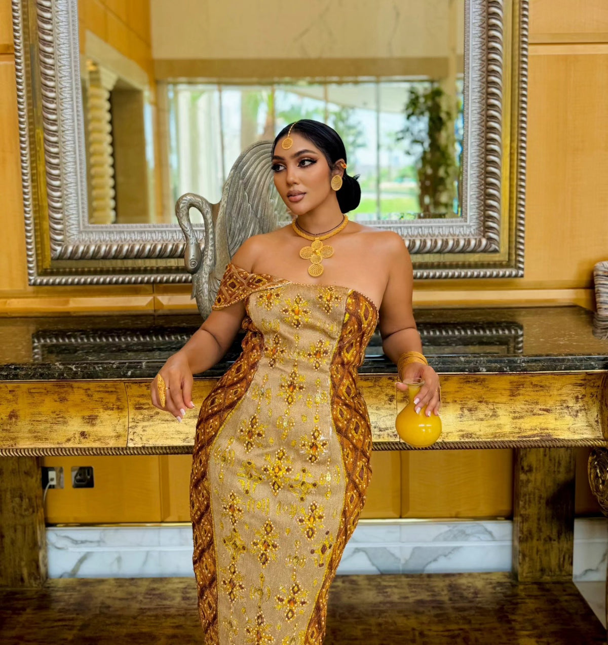 A woman stands indoors wearing a strapless beige and gold patterned habesha Wedding dress with brown draped side panels, posed in front of a large ornate mirror and wooden console.