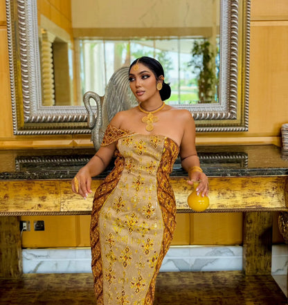 A woman stands indoors wearing a strapless beige and gold patterned habesha Wedding dress with brown draped side panels, posed in front of a large ornate mirror and wooden console.