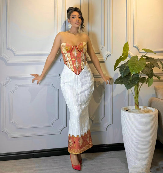 A woman models a traditional Thai-style strapless Habesha wedding dress featuring a red and gold embroidered corset bodice and a white pleated skirt with a matching red and gold patterned hem, standing in front of a white paneled wall.