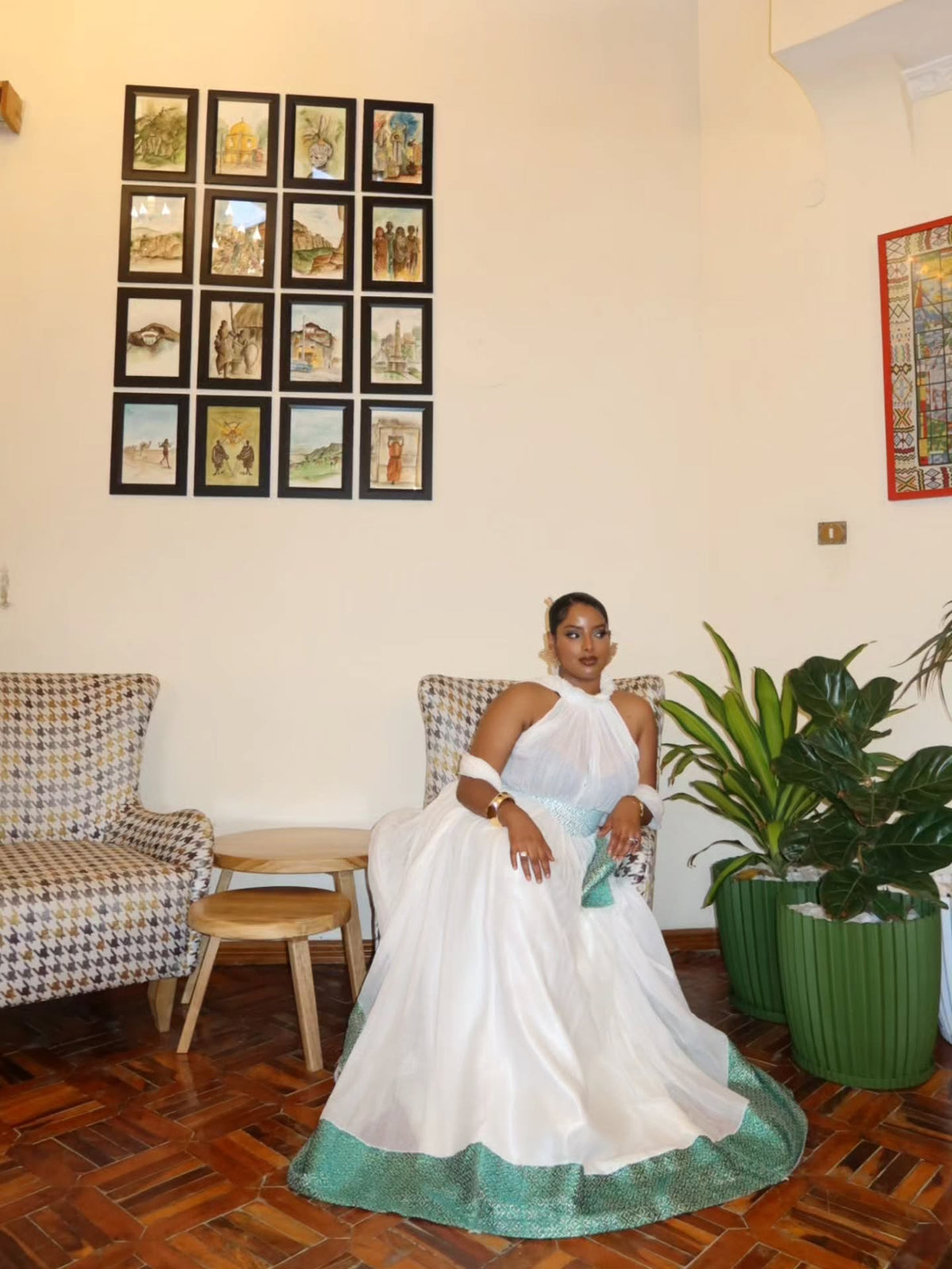 A woman wearing a white halter-neck ball Habesha Wedding Dress with a teal embroidered hem sits in a patterned armchair inside a home setting with wooden floors, potted plants, and framed pictures on the wall.