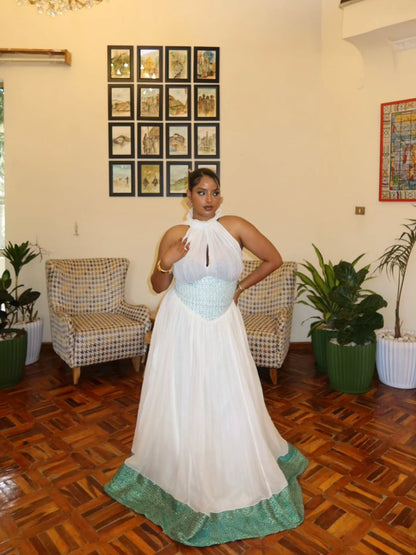 A woman modeling a white halter-neck maxi Habesha Wedding Dress with a light blue sequined bodice and a green lace-trimmed hem, standing in an indoor room with wooden floors, patterned armchairs, and potted plants.