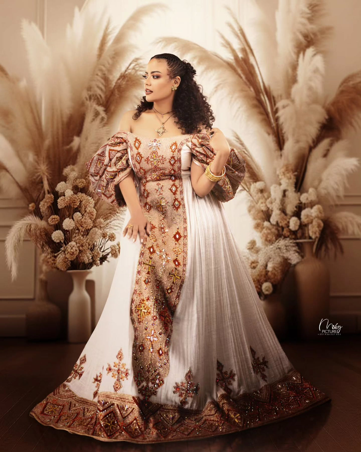 A woman models an ornate off-the-shoulder traditional Habesha Wedding Dress  with intricate gold, brown, and red embroidery on a white base, styled with a matching sheer white dupatta. She is posed in front of a warm, decorative background featuring pampas grass and dried floral arrangements.