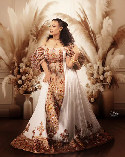 A woman models an ornate off-the-shoulder traditional Habesha Wedding Dress  with intricate gold, brown, and red embroidery on a white base, styled with a matching sheer white dupatta. She is posed in front of a warm, decorative background featuring pampas grass and dried floral arrangements.