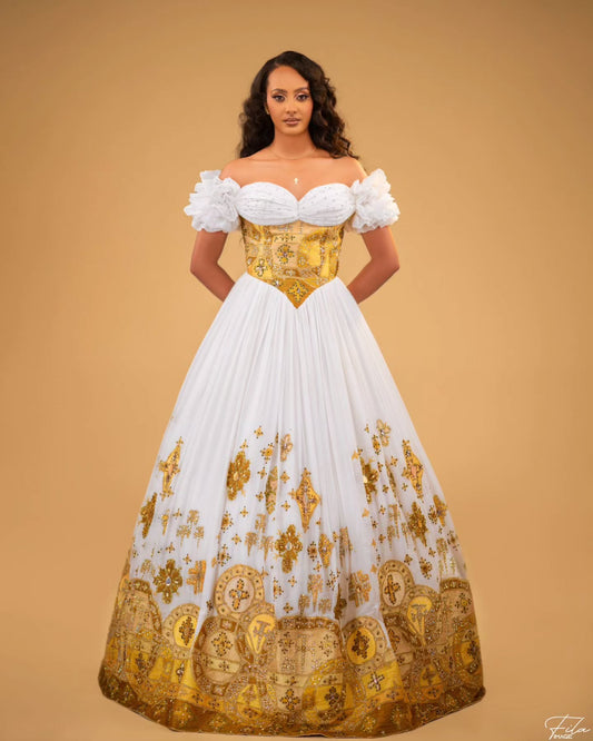 A woman modeling an off-the-shoulder ball Habesha Wedding Dress  with a white sweetheart bodice, puffed sleeves, and a full white skirt decorated with ornate gold baroque-style patterns, set against a solid tan background.