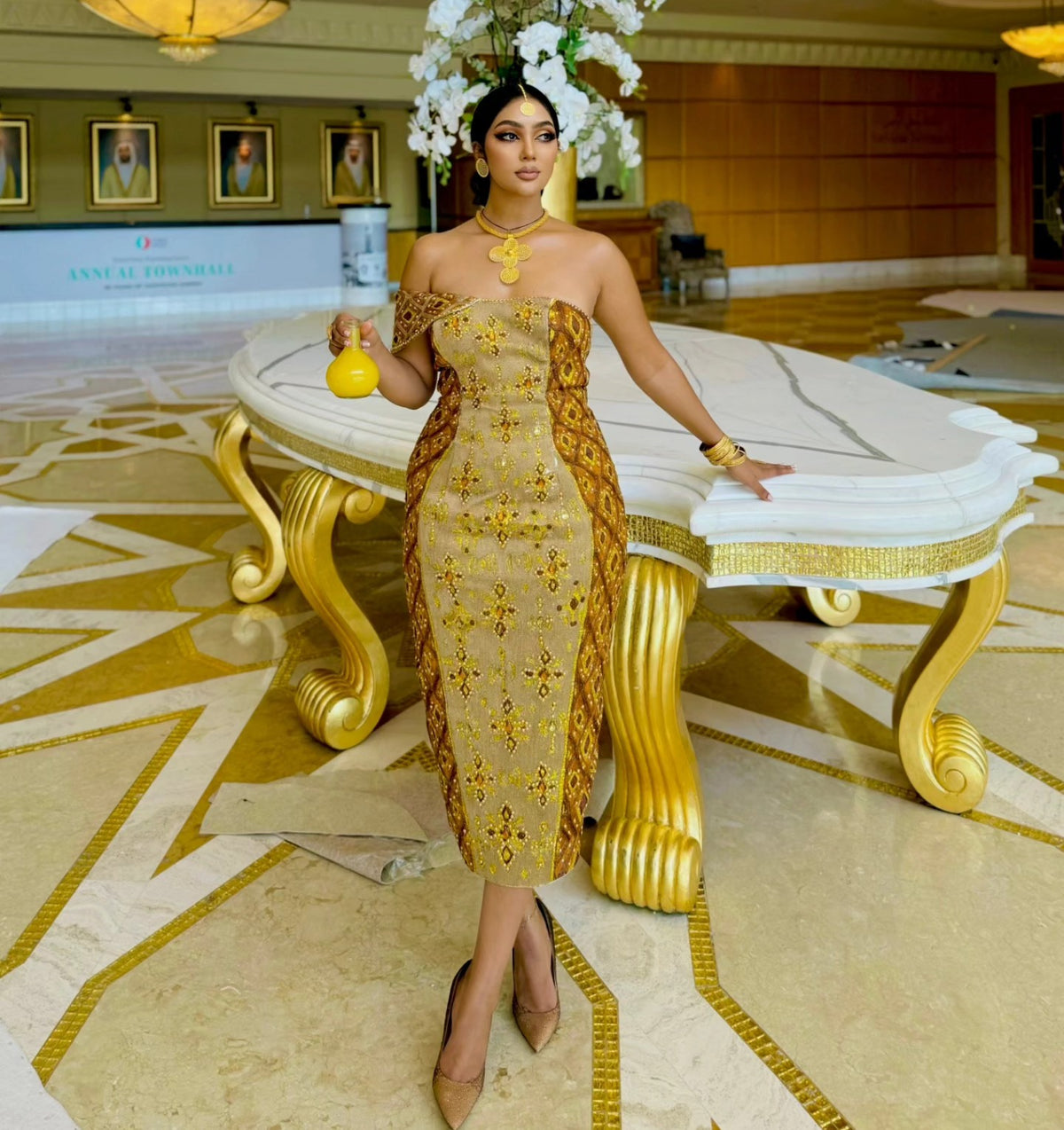A woman stands in a grand hotel lobby wearing a strapless beige and gold embroidered habesha Wedding dress with a matching headpiece of white flowers. She is holding a small yellow vase and posing in front of a large white and gold table.
