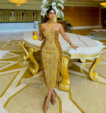 A woman stands in a grand hotel lobby wearing a strapless beige and gold embroidered habesha Wedding dress with a matching headpiece of white flowers. She is holding a small yellow vase and posing in front of a large white and gold table.