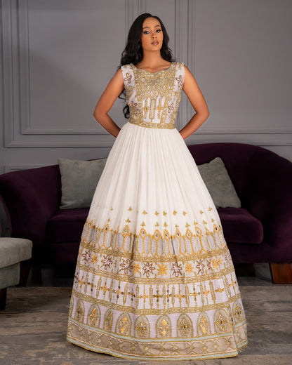 A woman modeling a sleeveless white and gold embellished ball Habesha Wedding Dress  in an indoor setting with a purple sofa in the background.