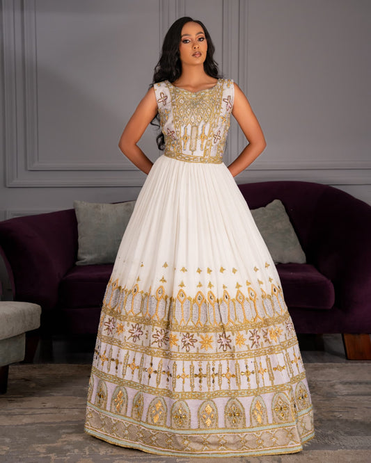 A woman modeling a sleeveless white and gold embellished ball Habesha Wedding Dress  in an indoor setting with a purple sofa in the background.