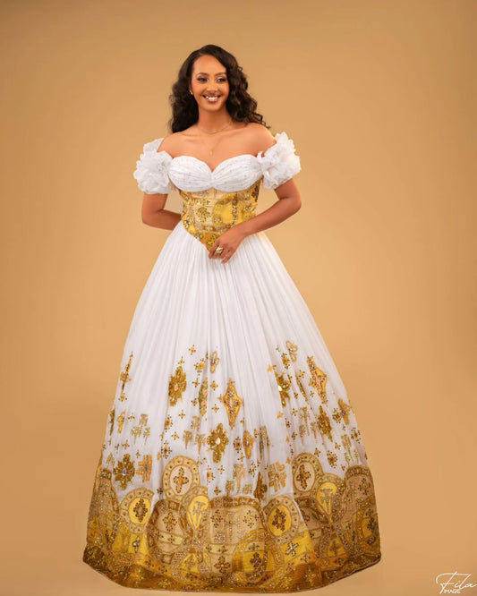 A woman modeling a white Habesha Wedding Dress with a structured gold corset bodice and ornate gold embroidery on the skirt, set against a solid tan studio background.
