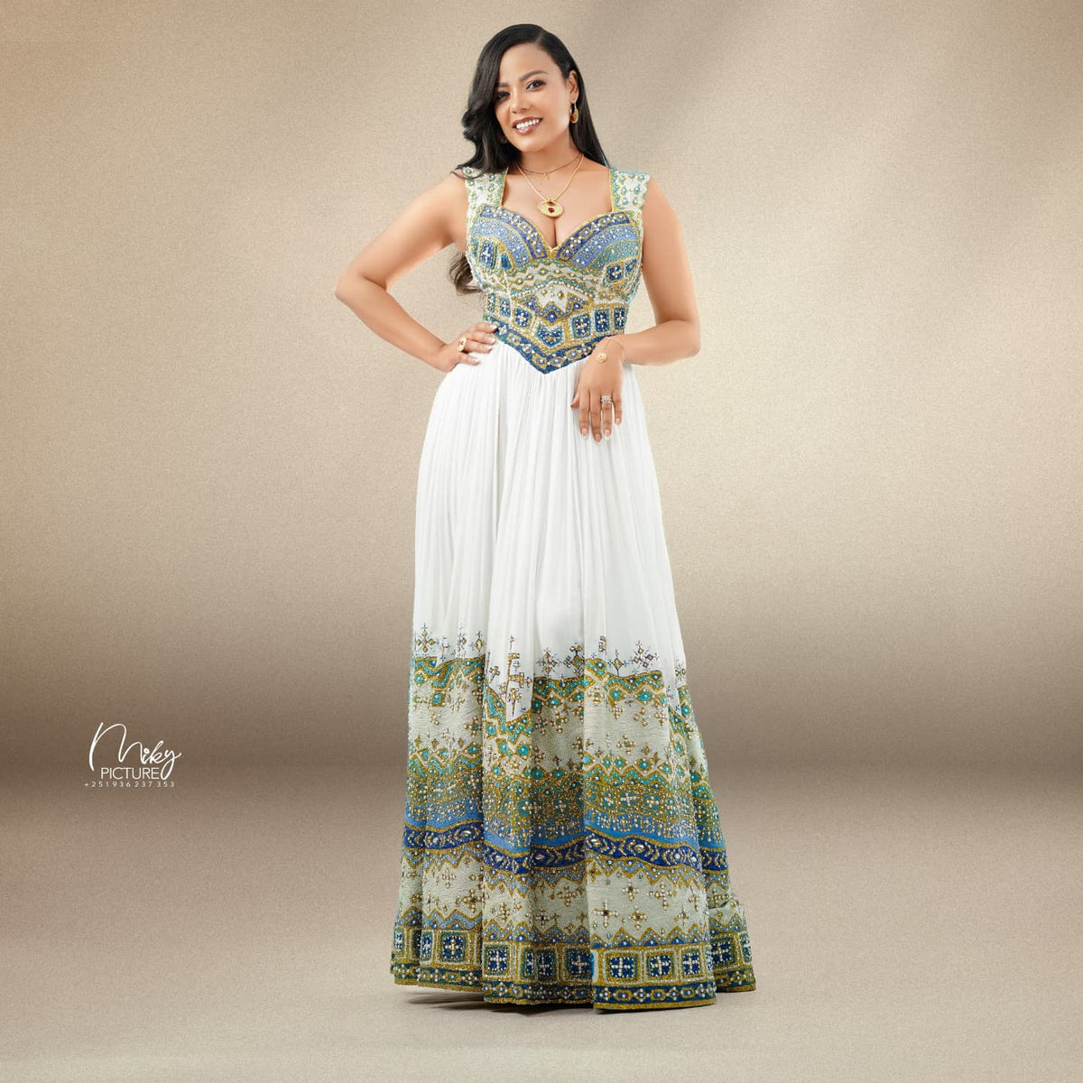 A woman modeling a sleeveless maxi habesha Wedding dress with a white pleated skirt and a heavily embroidered bodice and hem in blue, gold, and green patterns, set against a neutral beige studio background.