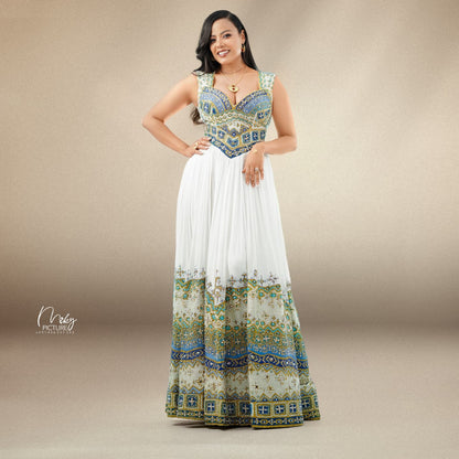 A woman modeling a sleeveless maxi habesha Wedding dress with a white pleated skirt and a heavily embroidered bodice and hem in blue, gold, and green patterns, set against a neutral beige studio background.