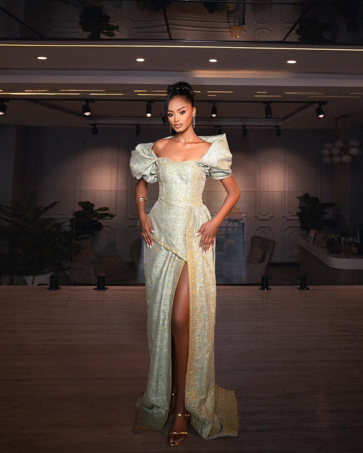 A woman stands in a dimly lit indoor venue wearing a formal off-the-shoulder Habesha Wedding Dress with a high thigh slit. The dress is a pale iridescent blue-green with gold patterning and a gold side panel, paired with metallic gold high-heeled sandals.