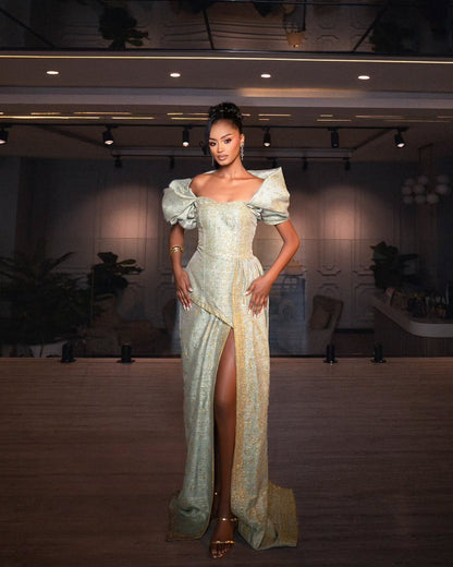 A woman stands in a dimly lit indoor venue wearing a formal off-the-shoulder Habesha Wedding Dress with a high thigh slit. The dress is a pale iridescent blue-green with gold patterning and a gold side panel, paired with metallic gold high-heeled sandals.