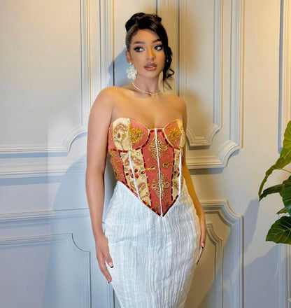 A woman models a strapless corset-style top with a red, gold, and beige patterned Habesha wedding dress, paired with a white pleated skirt, standing against a light blue paneled wall.