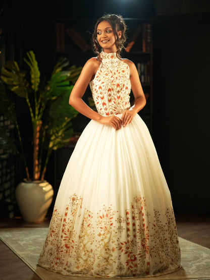 A woman modeling a sleeveless high-neck ball Habesha Wedding Dress  with a cream base and orange-brown floral embroidery concentrated on the bodice and lower skirt, posed in a dark indoor setting with a potted plant behind her.