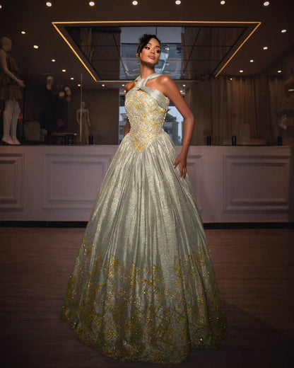 A woman modeling a floor-length halter ball Habesha Wedding Dress with a shimmering pale green body and gold floral embroidery, standing in a boutique interior.
