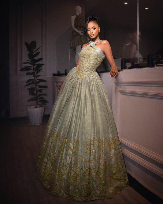 A woman models a floor-length ball Habesha Wedding Dress in a dimly lit indoor setting. The gown is a pale sage green with intricate gold embroidery concentrated on the bodice and lower skirt, featuring a halter neckline and a voluminous skirt.