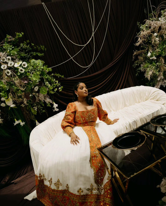 A woman wearing a traditional Habesha Wedding Dress  sits on a draped white sofa in an elegant setting with dark drapery, floral arrangements, and a glass table.