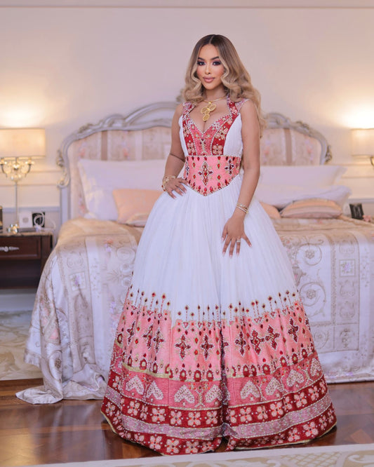 A woman models a traditional South Asian ball habesha Wedding dress in a bedroom setting. The dress features a white tulle skirt with a heavily embroidered pink and red border, a pink and red embroidered bodice with white draped shoulder panels, and a gold chain necklace.